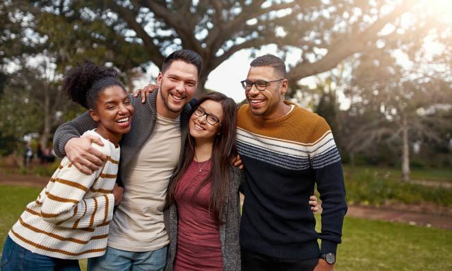 Four young adult friends smiling with arms around each other outside in a park
