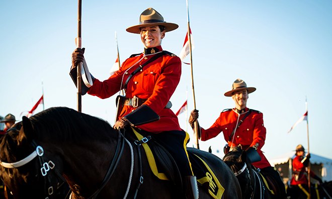 A woman and man are shown holding flags while riding horses, with a clear blue sky in the background. Both are wearing traditional red RCMP uniforms.