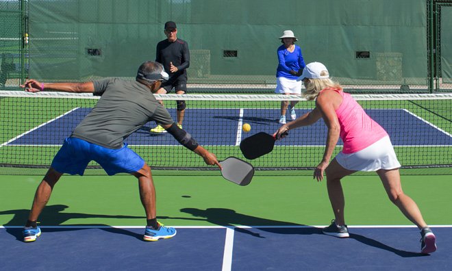 Four people playing a game of pickleball in an outdoor court. A man and woman on the same team are both lunging to hit a ball that has just passed over the net.
