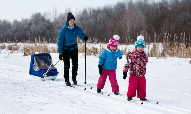 Mother enjoying a cross-country ski trail with three daughters. She is pulling the smallest daughter in a sled behind her.