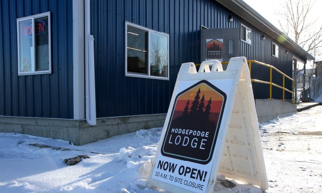 Image of a building with blue siding, there is a concrete ramp with yellow railing leading up to an open door. A sign in the foreground says HodgePodge Lodge Now Open