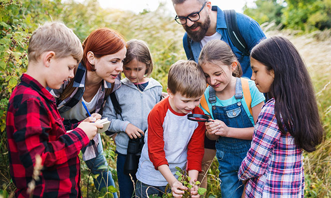 A small group of kids and two adults outdoors, looking at a plant through a magnifying glass
