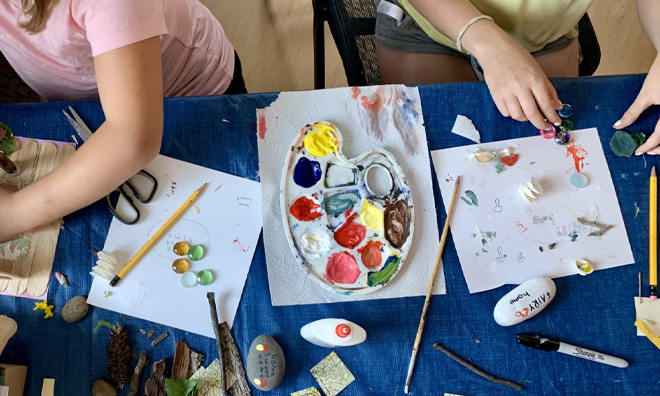 Overhead shot of two young girls working on a blue table with an assortment of paintings and painting tools.