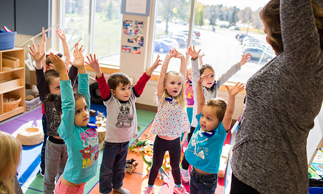 Group of young children raising their hands in their air while looking at a preschool instructor