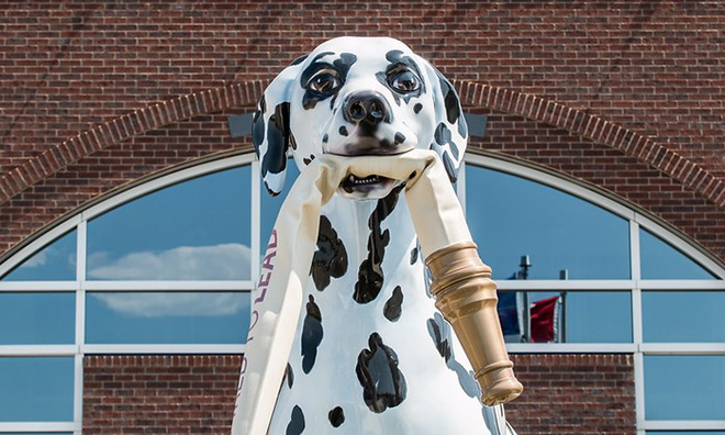 Close up of a large sculpture of a dalmatian dog holding the end of a fire hose in its mouth.