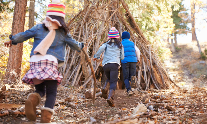 Three children shown running away from the camera towards a tree fort. All three are dressed in coats in wearing toques.