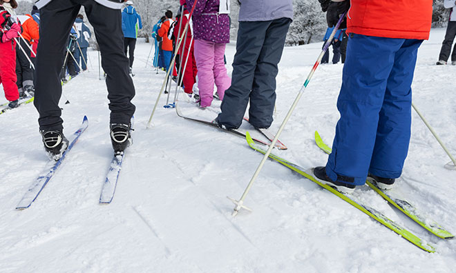 Bottom half of various skiiers of all ages are shown waiting in a lineup in the snow. All are wearing snowpants and cross-country skiis.