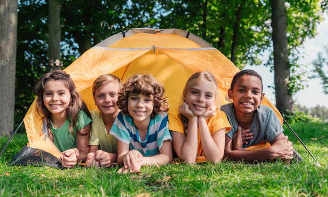 Several children are smiling towards the camera while laying down in a forested area. Their feet are extending towards a small orange tent in the background.