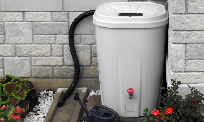 Image of a rain barrel sitting next to the side of a house. There are flowers in a stone covered flower bed at the base of the rain barrel
