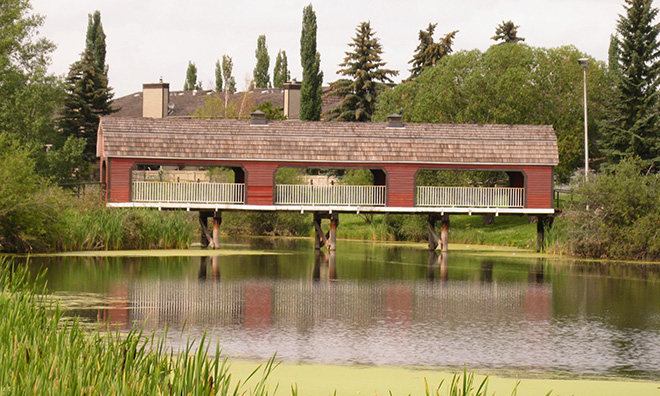 Image of a body of water surrounded by cattails and trees with a wooden covered bridge in the background.