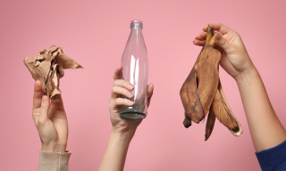 Image of three hands on a pink background, the hands are holding a piece of crumpled paper, and empty glass bottle, and a banana skin.